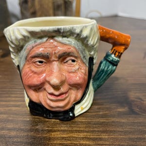 May include: A ceramic mug shaped like an elderly woman's face. The mug features a detailed painted face with rosy cheeks, a white bonnet, and a black bow. The handle is orange and green.