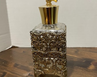 May include: A vintage perfume bottle with a clear glass body and a decorative gold-colored metal filigree cage. The bottle has a gold-colored cap and atomizer. The perfume bottle is rectangular and sits on four small feet.
