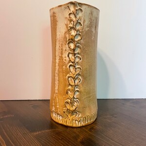 May include: A ceramic vase with a light brown glaze and a white design of leaves running vertically down the side.