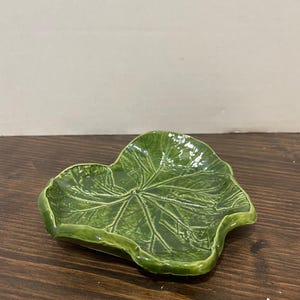 Puede incluir: Un plato de cerámica verde oscuro brillante con forma de hoja grande similar a un corazón. La superficie presenta una textura detallada y en relieve que imita las venas naturales de las hojas, con un borde ligeramente ondulado e irregular.