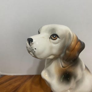 May include: A ceramic dog figurine featuring a white base coat with brown and black patches on the ears and body. The dog has painted brown eyes, a black nose, and small black spots on the muzzle, captured in a seated position facing left.