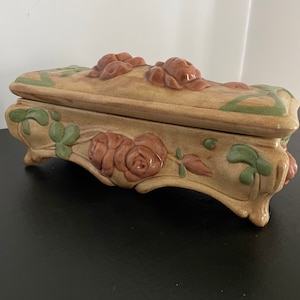 May include: A ceramic trinket box with a floral design. The box is decorated with raised roses and leaves in shades of brown, green, and beige. The box has a rectangular shape with a slightly curved top and four small feet.