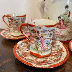 May include: A porcelain tea set featuring two cups and saucers with a white base, orange rims, and hand-painted scenes of figures in traditional clothing, trees, and buildings. The set includes a matching teapot with similar decorative motifs.