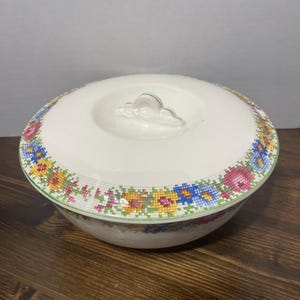 Könnte beinhalten: Eine weiße Keramik-Servierschüssel mit Deckel. Schüssel und Deckel sind mit einem floralen Muster in Grün, Gelb, Blau und Rosa verziert.