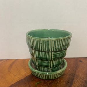 Pode incluir: Um vaso de plantas em cerâmica verde com pires a condizer, apresentando um exterior texturizado com padrões de nervuras verticais e horizontais alternadas. O esmalte tem um acabamento brilhante e o vaso tem um topo largo e aberto.