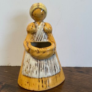 Puede incluir: Una figurita de cerámica de una mujer con un vestido amarillo y un delantal blanco que sostiene una cesta. La figurita es una maceta con un agujero en la cesta para plantar.