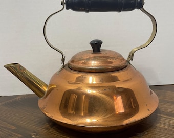 May include: A copper-colored kettle with a black wooden handle and a brass spout. The kettle has a round body and a lid with a small black knob. The handle is attached to the kettle with metal brackets.
