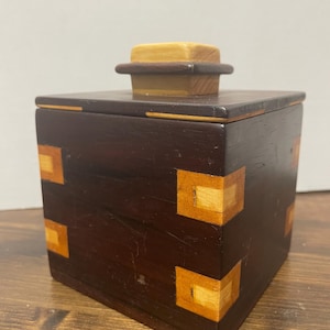 May include: A dark brown wooden box with a square shape and a light-colored wooden lid. The corners of the box are decorated with square inlays of light-colored wood. The box is sitting on a wooden surface.