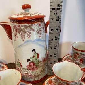 May include: A porcelain tea set featuring a tall teapot with a lid and matching cups on saucers, decorated with hand-painted figures in traditional attire and floral motifs. A metal ruler shows the teapot height is approximately 8.5 inches.