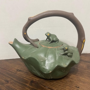 Puede incluir: Una tetera de color verde mate con forma de nenúfar grande con dos pequeñas ranas verdes posadas en la tapa y en el lateral. El asa es marrón y tiene una textura que recuerda a una rama de árbol, y el pico está integrado en el diseño de hoja.