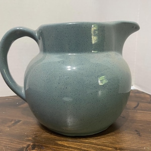May include: A speckled, blue-green ceramic pitcher with a rounded body, a curved handle, and a pouring spout. The pitcher is sitting on a wooden surface.