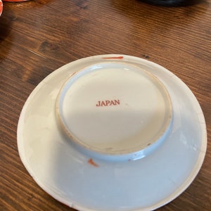May include: A white ceramic saucer viewed from the underside, featuring a circular foot rim and the word JAPAN printed in red ink at the center. The edge of the saucer shows a thin orange-red painted border with minor signs of wear and aging.