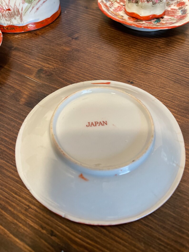 May include: A white ceramic saucer viewed from the underside, featuring a circular foot rim and the word JAPAN printed in red ink at the center. The edge of the saucer shows a thin orange-red painted border with minor signs of wear and aging.
