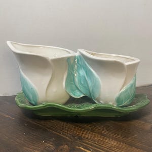 Peut inclure: Une jardinière double en céramique composée de deux récipients en forme de tulipe de couleur crème avec des feuilles turquoise clair sur les côtés. Les deux récipients sont reliés et reposent sur un plateau en céramique vert foncé.