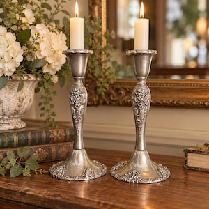 May include: Two silver-toned metal candlesticks with ornate embossed floral and grape vine detailing on the stems and bases. Each candlestick holds a single tall, cylindrical white wax candle with a lit flame at the top.