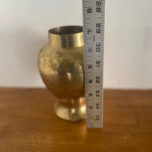 May include: A gold-colored brass vase with a rounded body and a flared lip. The vase is approximately 9 inches tall and is sitting on a wooden surface.