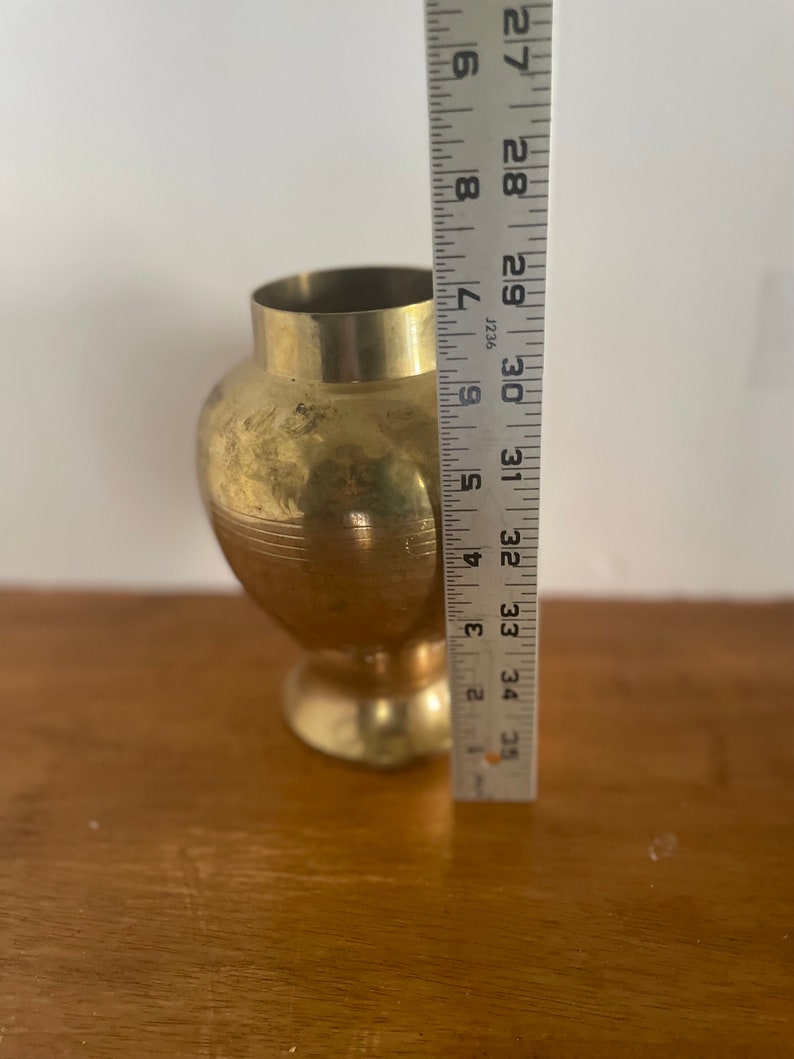 May include: A gold-colored brass vase with a rounded body and a flared lip. The vase is approximately 9 inches tall and is sitting on a wooden surface.