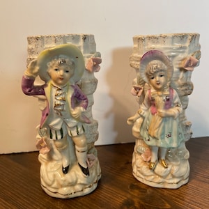 May include: Pair of vintage porcelain vases featuring a boy and girl in 18th century attire. The vases are decorated with gold accents and floral details.