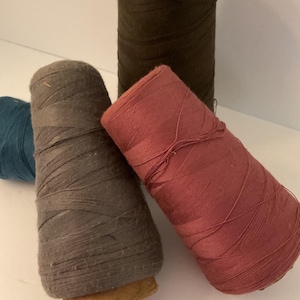 May include: Four spools of thread in different colors: dark brown, gray, pink, and blue. The spools are made of cardboard and have a wooden base.