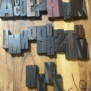 May include: A collection of vintage wooden letterpress blocks featuring the full alphabet from A to Z, arranged in three rows. The blocks are dark brown with visible wood grain and show signs of wear. A handwritten hashtag and number 6 are visible.