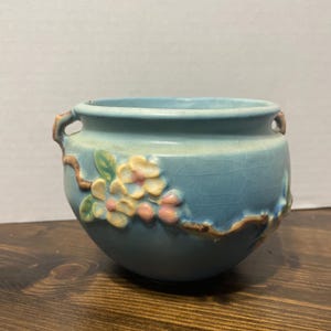 May include: A light blue ceramic planter with a floral design. The planter has a round shape with two small handles and a textured surface. The floral design features pink and yellow flowers with green leaves. The planter is sitting on a wooden surface.