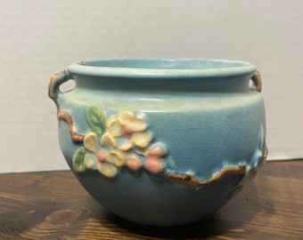 May include: A light blue ceramic planter with a floral design. The planter has a round shape with two small handles and a textured surface. The floral design features pink and yellow flowers with green leaves. The planter is sitting on a wooden surface.