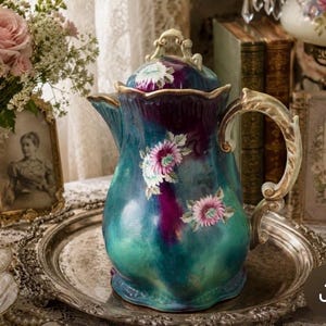 May include: A ceramic coffee pot featuring a deep teal and purple gradient glaze with hand-painted pink and white floral accents. The pot has a scalloped rim, a curved handle with gold-toned detailing, and a matching lid with a decorative finial.