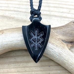 May include: A black shield-shaped pendant with an intricate engraved design, suspended from a black braided cord. The pendant is made of a dark, possibly obsidian-like material. The background features a wooden surface and a light-colored branch.