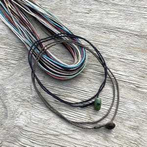 Puede incluir: Collares de cordón trenzado en varios colores, incluyendo negro, marrón y hebras multicolores. Un collar presenta un colgante de piedra verde, y otro tiene una pequeña cuenta marrón oscuro. Los cordones se muestran sobre una superficie de madera envejecida.