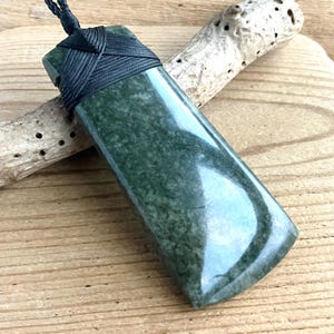 May include: A rectangular pendant made of polished dark green stone with a mottled texture. The top of the pendant is secured with a black braided cord that wraps around the stone in a crisscross pattern, forming a loop for a necklace.
