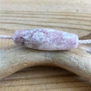Peut inclure: Une perle cylindrique en forme de tonneau avec une surface texturée et marbrée dans des tons de rose pâle, de blanc et de lavande doux. La perle est enfilée sur un cordon tressé fin et clair, présentant un aspect naturel rappelant la pierre.