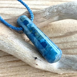 Könnte beinhalten: Zylindrischer Anhänger in Blau- und Türkistönen, an einer geflochtenen blauen Kordel. Der Anhänger hat einen marmorierten Effekt. Die Kordel ist oben am Anhänger befestigt.