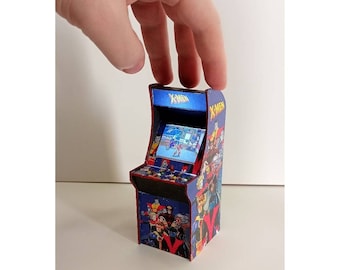 Puede incluir: Mini máquina recreativa con temática de X-Men. La máquina es principalmente azul con detalles rojos y muestra una pantalla de juego. Los lados y la parte superior están decorados con arte de cómics de X-Men. Una mano sostiene la máquina.