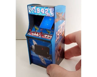 Puede incluir: Miniatura de máquina recreativa con exterior azul, con el título del juego "1942" e imágenes de aviones de combate. La máquina tiene joysticks rojos y un panel de control detallado, evocando un tema de juego retro.