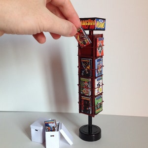 Peut inclure: Présentoir miniature avec des magazines de jeux vidéo. Le présentoir est rouge foncé avec une base noire et porte le texte "GamePro Visions" en haut. Plusieurs magazines sont visibles, et une main en tient un.