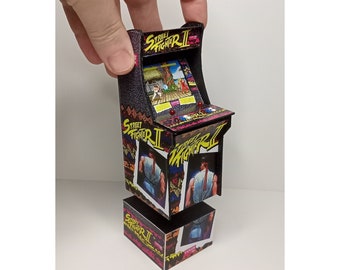 Puede incluir: Una réplica de arcade en miniatura del clásico videojuego Street Fighter II. El gabinete es negro con un diseño rojo y amarillo y presenta el logotipo y los personajes del juego.