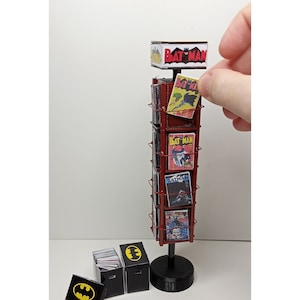 Op de afbeelding: Een miniatuur stripboek displaystandaard met Batman strips. De standaard is rood met een zwarte basis en bevat verschillende stripboeken. Er zijn ook kleine zwarte dozen met het Batman-logo en een Batman-logo op een kleine zwarte rechthoek.