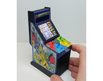 Könnte beinhalten: Miniatur-Spielautomat mit dem Spiel "Gauntlet". Der Automat ist schwarz mit farbenfrohen Grafiken an den Seiten und einem Bildschirm, der das Gameplay anzeigt. Er hat Joysticks und Knöpfe in verschiedenen Farben. Oben steht "Gauntlet" in Blau.