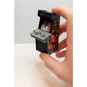 May include: Miniature arcade cabinet with a black base and colorful side panels featuring game characters. The control panel has small joysticks and buttons. The screen displays a game scene. The cabinet is held in a hand.