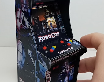 May include: Miniature Robocop arcade cabinet with a black exterior. The cabinet features the Robocop logo and imagery, with red, yellow, and blue buttons. The screen displays a game scene. The cabinet is held by a hand.