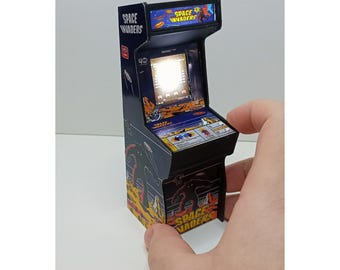 Puede incluir: Mini máquina recreativa de Space Invaders. El mueble es negro con gráficos coloridos y el logotipo del juego. La pantalla muestra la interfaz del juego. La máquina recreativa está sujeta por una mano.