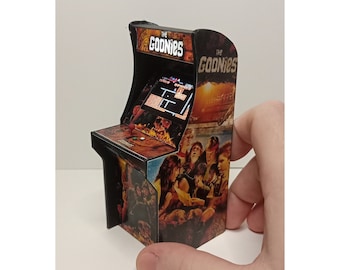 Puede incluir: Miniatura de máquina recreativa con el texto "THE GOONIES" en la parte superior. El mueble tiene una pantalla que muestra un videojuego pixelado y está decorado con imágenes de la película. El mueble está sostenido por una mano.