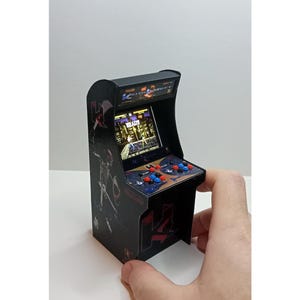 May include: Miniature arcade cabinet with a black exterior, featuring a screen displaying a game interface. The cabinet has red, blue, and black buttons and a joystick. The text "READY" is visible on the screen. The cabinet is held by a hand.