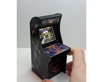 Puede incluir: Mini máquina recreativa con exterior negro, con una pantalla que muestra una interfaz de juego. La máquina tiene botones rojos, azules y negros y un joystick. El texto "READY" es visible en la pantalla. La máquina es sostenida por una mano.