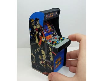 Puede incluir: Miniatura de máquina recreativa con parte superior negra y laterales azules con gráficos de juegos de acción. El mueble tiene un joystick y botones, y está sostenido por una mano. Un objeto de colección de juegos retro.