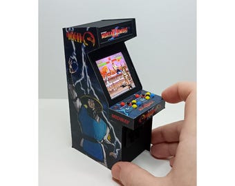 Puede incluir: Miniatura de máquina recreativa Mortal Kombat con el logotipo del juego e ilustraciones de personajes. El mueble es negro, con una pantalla que muestra el juego y botones coloridos. El logotipo de Midway es visible en la parte frontal.