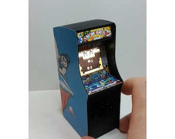 Puede incluir: Miniatura de máquina recreativa con el juego "Snow Bros.". El mueble tiene un diseño azul y negro con gráficos del juego. La pantalla muestra una escena de juego pixelada. El mueble es sostenido por una mano.