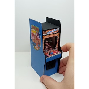 May include: A miniature blue arcade cabinet featuring the Donkey Kong game. The cabinet has a black top and a colorful screen displaying the game's graphics. The side panel shows the game's logo. The cabinet is held by a hand.