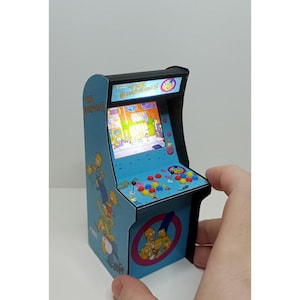 Puede incluir: Una máquina recreativa en miniatura con el tema de "Los Simpson". El mueble azul claro tiene una pantalla que muestra un juego y botones coloridos. Los laterales están decorados con el logotipo y los personajes de la serie.