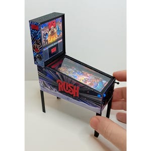 Könnte beinhalten: Miniatur-Flipperautomat mit dem Wort "RUSH" in Rot. Die Maschine hat einen schwarzen Rahmen und Beine, mit einem farbenfrohen Spielfeld unter Glas sichtbar. Die Rückwand zeigt eine Grafik mit dem Wort "RUSH".